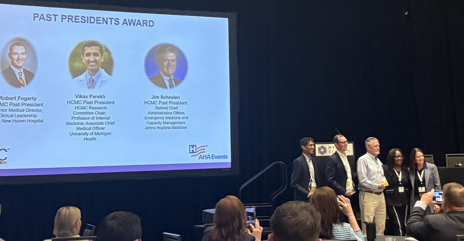 A group of people stands on stage receiving an award, with a screen behind them displaying the names and photos of past presidents.
