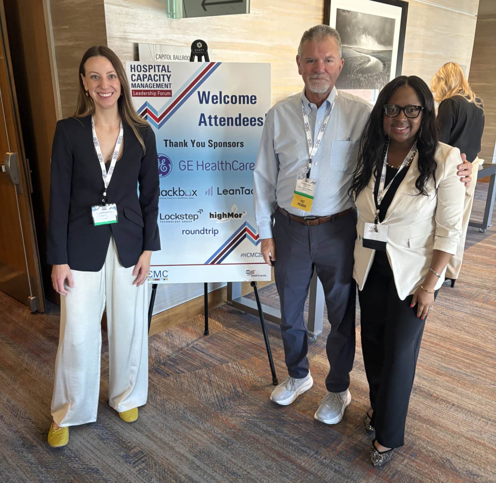 Three people stand smiling in front of a conference welcome sign for the Hospital Capacity Management Leadership Forum.