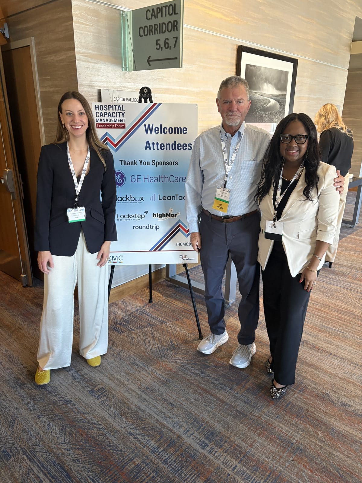Three people pose and smile beside a “Welcome Attendees” sign at a hospital capacity event, standing in a carpeted hallway with conference badges visible.