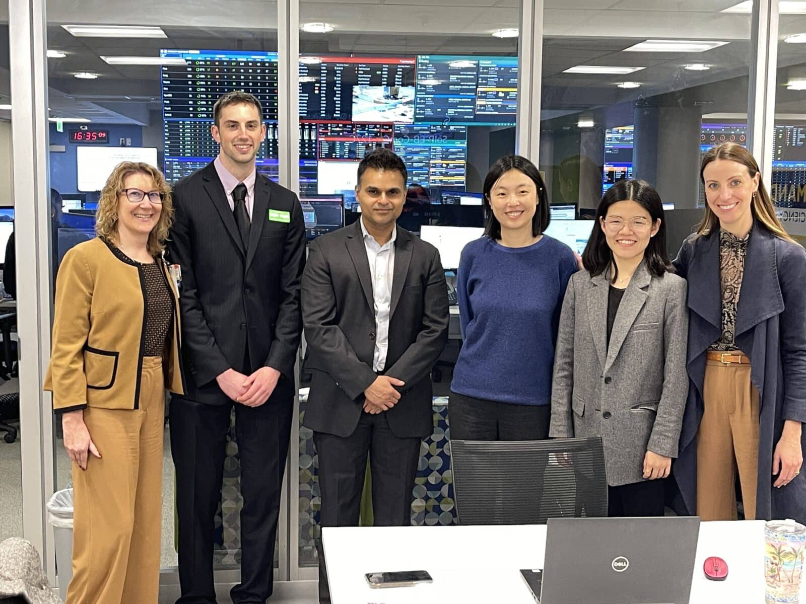 Six people in business attire stand together in an office with large monitors displaying data on the wall behind them.